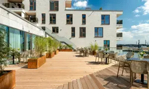 Holzterrasse mit Tischen, Stühlen und Pflanzen vor einem modernen Gebäude mit Blick auf den Hafen.