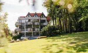 Hotelgebäude mit Balkonen und Spitzdächern, umgeben von Bäumen und einer grünen Wiese.