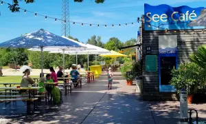 See Café mit Tischen und Gästen unter Sonnenschirmen am See.