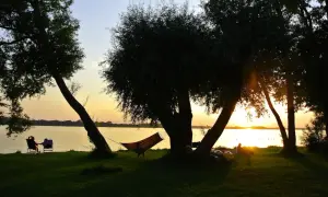 Sonnenuntergang am See mit Hängematte und Liegestühlen im Schatten von Bäumen.