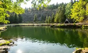Grüner See mit Bäumen, Felsen und einem kleinen Unterstand am Ufer.