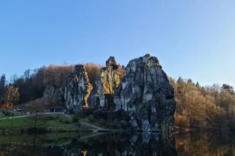 Felsformationen am Seeufer mit Spiegelung im Wasser und bewaldetem Hintergrund.
