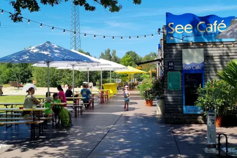 See Café mit Tischen und Gästen unter Sonnenschirmen am See.