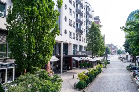 Das Madison Hotel Hamburg mit seinem Eingangsbereich und Außenbereich, umgeben von Bäumen und einer Kopfsteinpflasterstraße.