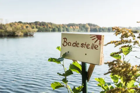 Schild "Badestelle" am See mit Bäumen im Hintergrund.