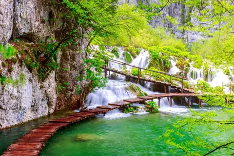 Holzstege über türkisfarbenes Wasser führen zu mehreren Wasserfällen im Grünen.