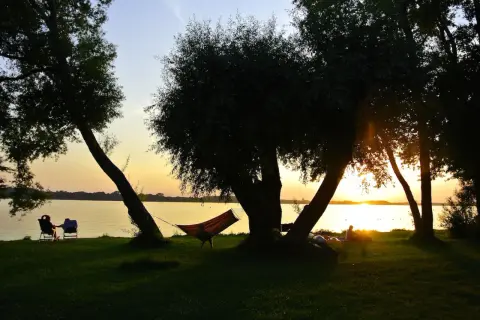 Sonnenuntergang am See mit Hängematte und Liegestühlen im Schatten von Bäumen.