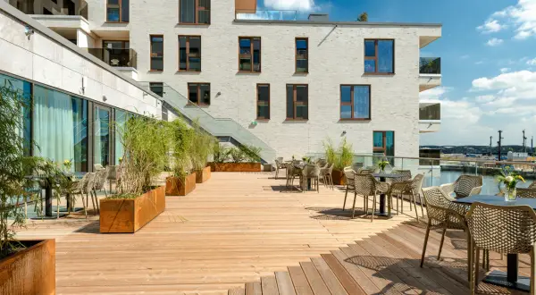 Holzterrasse mit Tischen, Stühlen und Pflanzen vor einem modernen Gebäude mit Blick auf den Hafen.
