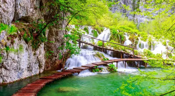 Holzstege über türkisfarbenes Wasser führen zu mehreren Wasserfällen im Grünen.