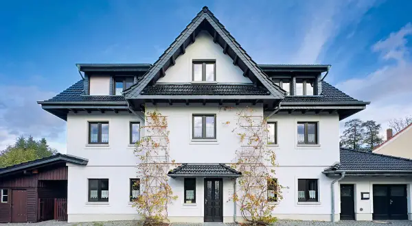 Weißes Haus mit dunklem Dach und Klinker-Vorplatz, bewachsen mit Weinreben.