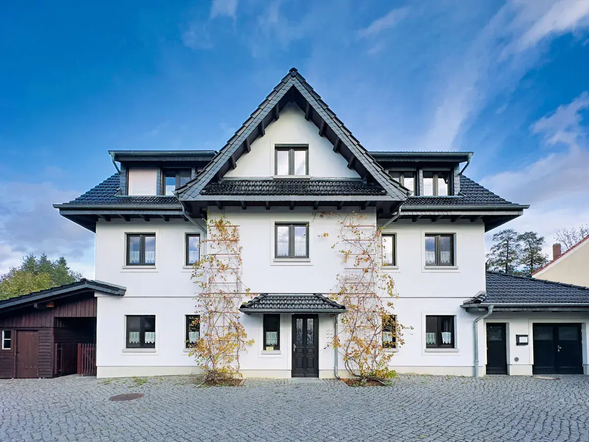 Weißes Haus mit dunklem Dach und Klinker-Vorplatz, bewachsen mit Weinreben.