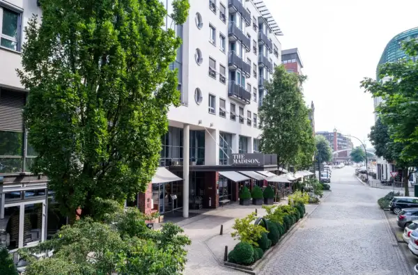 Das Madison Hotel Hamburg mit seinem Eingangsbereich und Außenbereich, umgeben von Bäumen und einer Kopfsteinpflasterstraße.