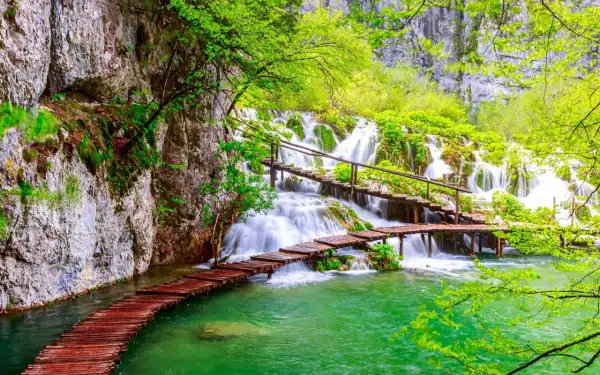 Holzstege über türkisfarbenes Wasser führen zu mehreren Wasserfällen im Grünen.