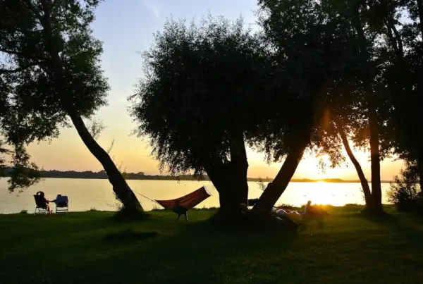 Sonnenuntergang am See mit Hängematte und Liegestühlen im Schatten von Bäumen.